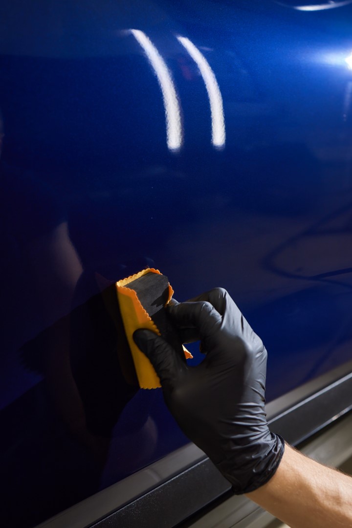 The process of applying a nano-ceramic coating on the white car's hood by a male worker in blue gloves with a sponge and chemical composition to protect the paint in detailing workshop. Auto service.