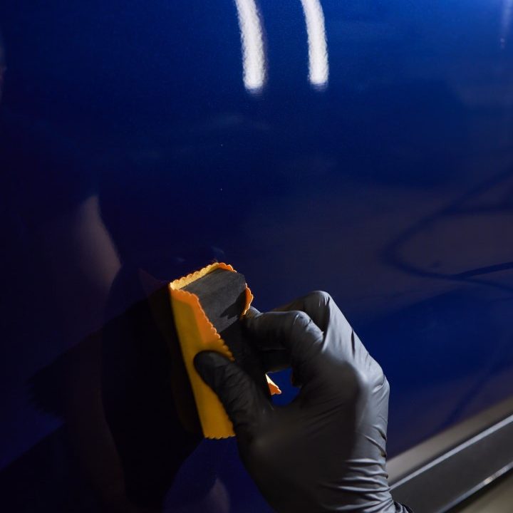 The process of applying a nano-ceramic coating on the white car's hood by a male worker in blue gloves with a sponge and chemical composition to protect the paint in detailing workshop. Auto service.